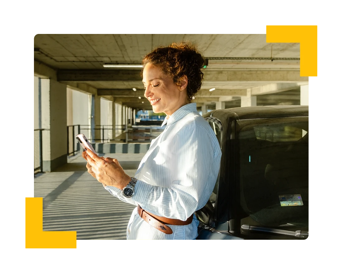 Resident smiling at her phone while leaning on her car in a parking garage